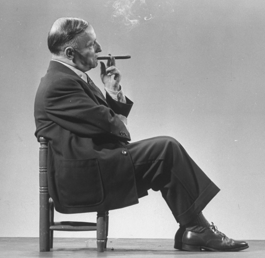 Architect Frederick Kiesler seated in a child's chair, smoking a cigar, at Gjon Mili's studio.